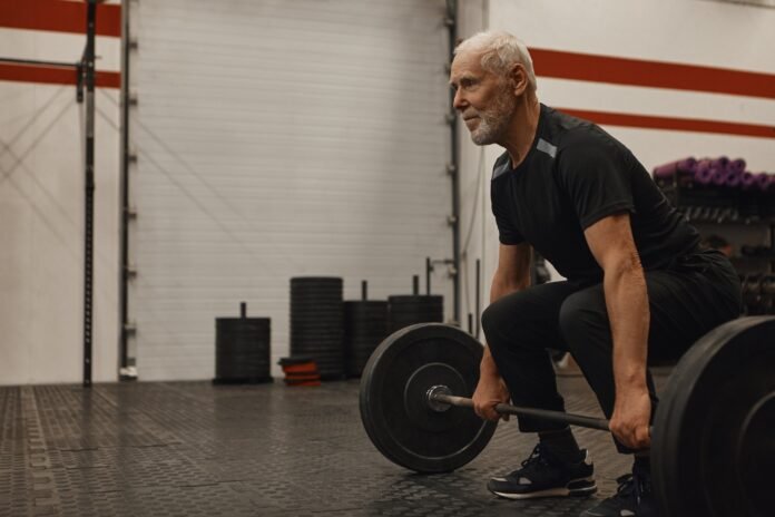 Stylish seventy year old male choosing healthy active lifestyle, training with weights indoors, lifting barbell, bulding muscular strong arms, having joufyl happy facial expression. Age and vitality